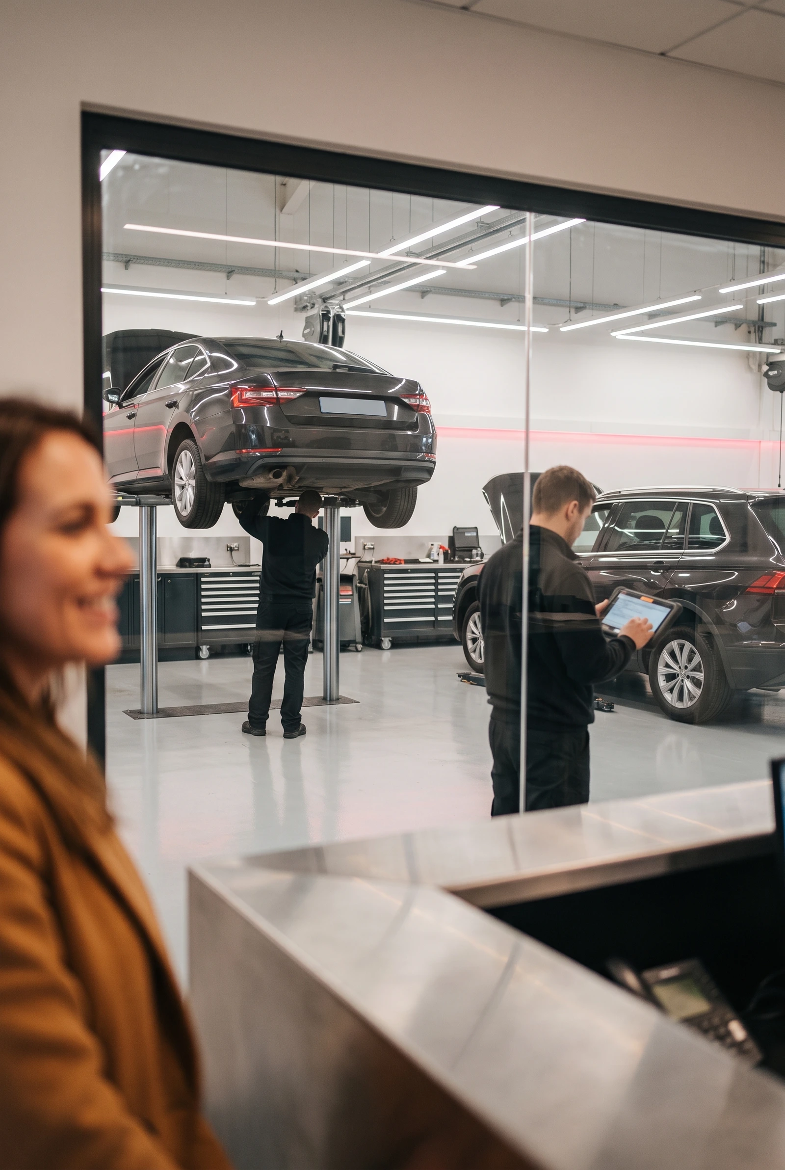 Technicians working on vehicles raised on lifts in a busy automotive workshop, seen through the reception glass partition